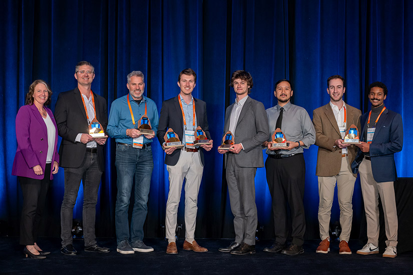 Several people stand on a stage posing for the camera, many of them holding awards.