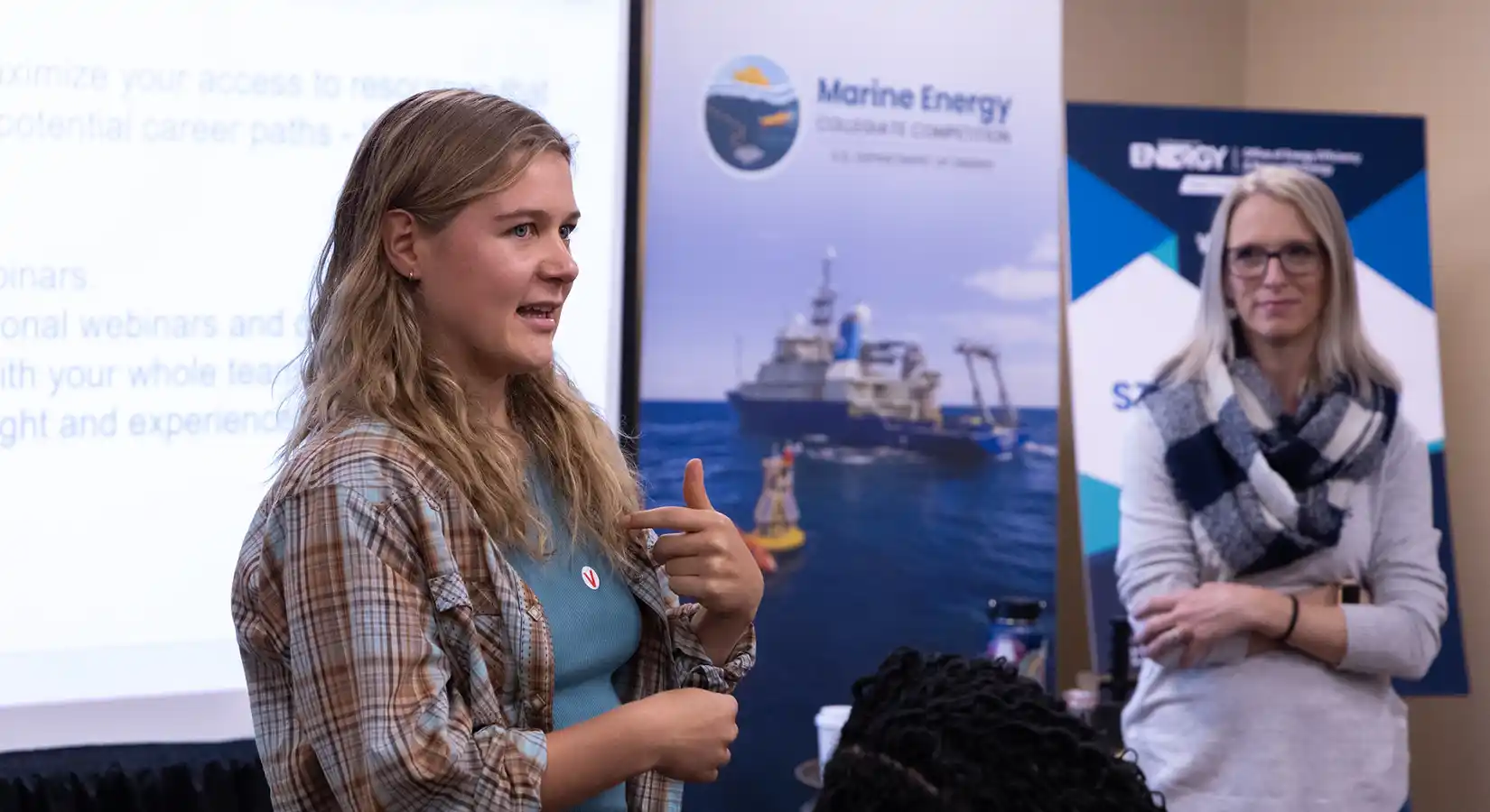 A person speaks in front of posters and a presentation screen as another person watches.