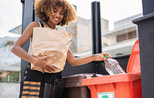 A woman holding a paper bag places plastic bottles into a recycling bin