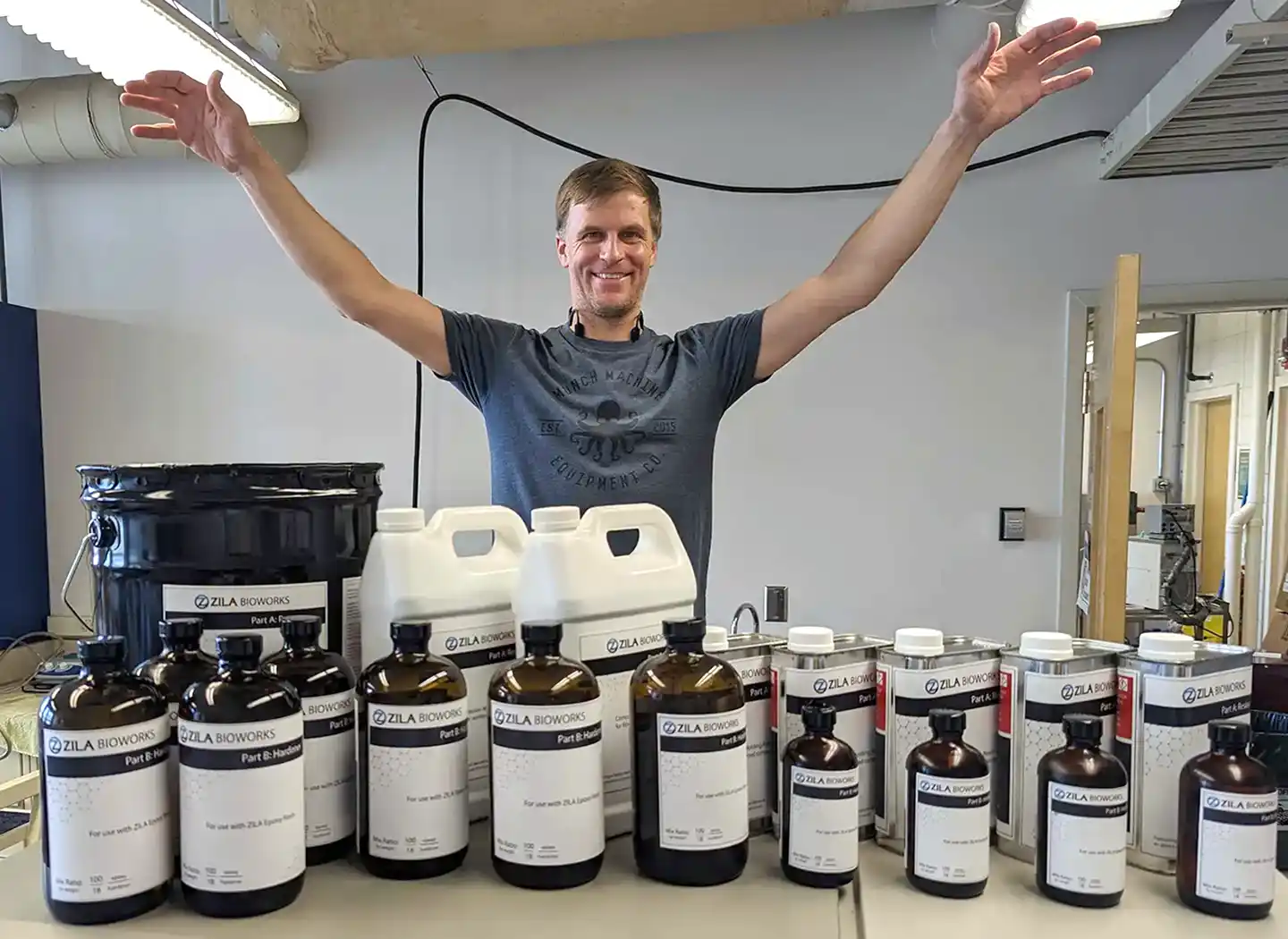 A researcher raises his arms, palms down, over a collection of containers with the name, “Zila Bioworks” written on them.