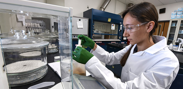 A woman in safety glasses, rubber gloves, and lab coat measures a sample in a laboratory.