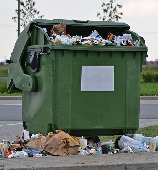 Trash bin in a parking lot overflowing with food packaging waste.