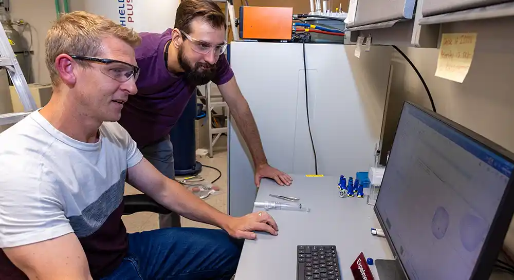 Two people in safety glasses look at a computer monitor.