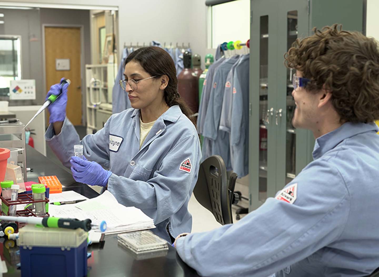 Two people in blue lab coats in a lab. One of them, a woman, is collecting material to put into a plastic tube.