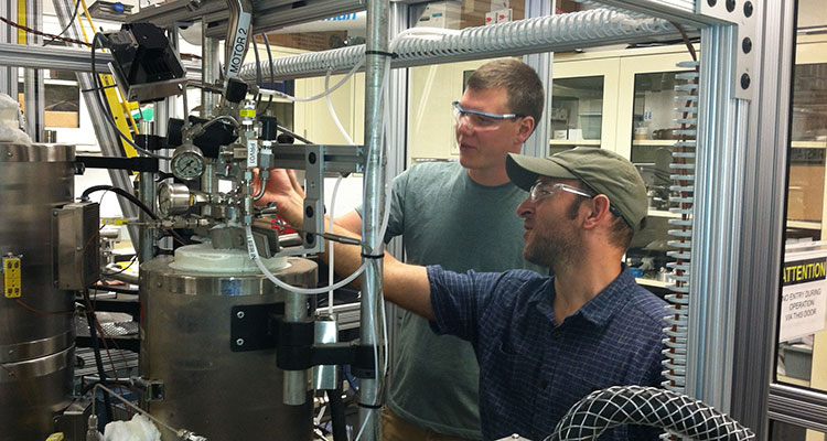 Photo of two engineers in safety glasses working on a reactor system with metal tanks, tubing, pipes, and wires. The system is used for in situ and ex situ catalytic fast pyrolysis to generate bio-oil for compositional analysis.