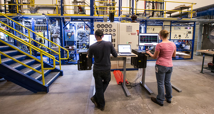 Photo of two engineers facing a large, multi-level Davison circulating riser, with laptops on tall tables. The riser features colorful scaffolding, pipes, tubes, and control panels with dials and monitors.