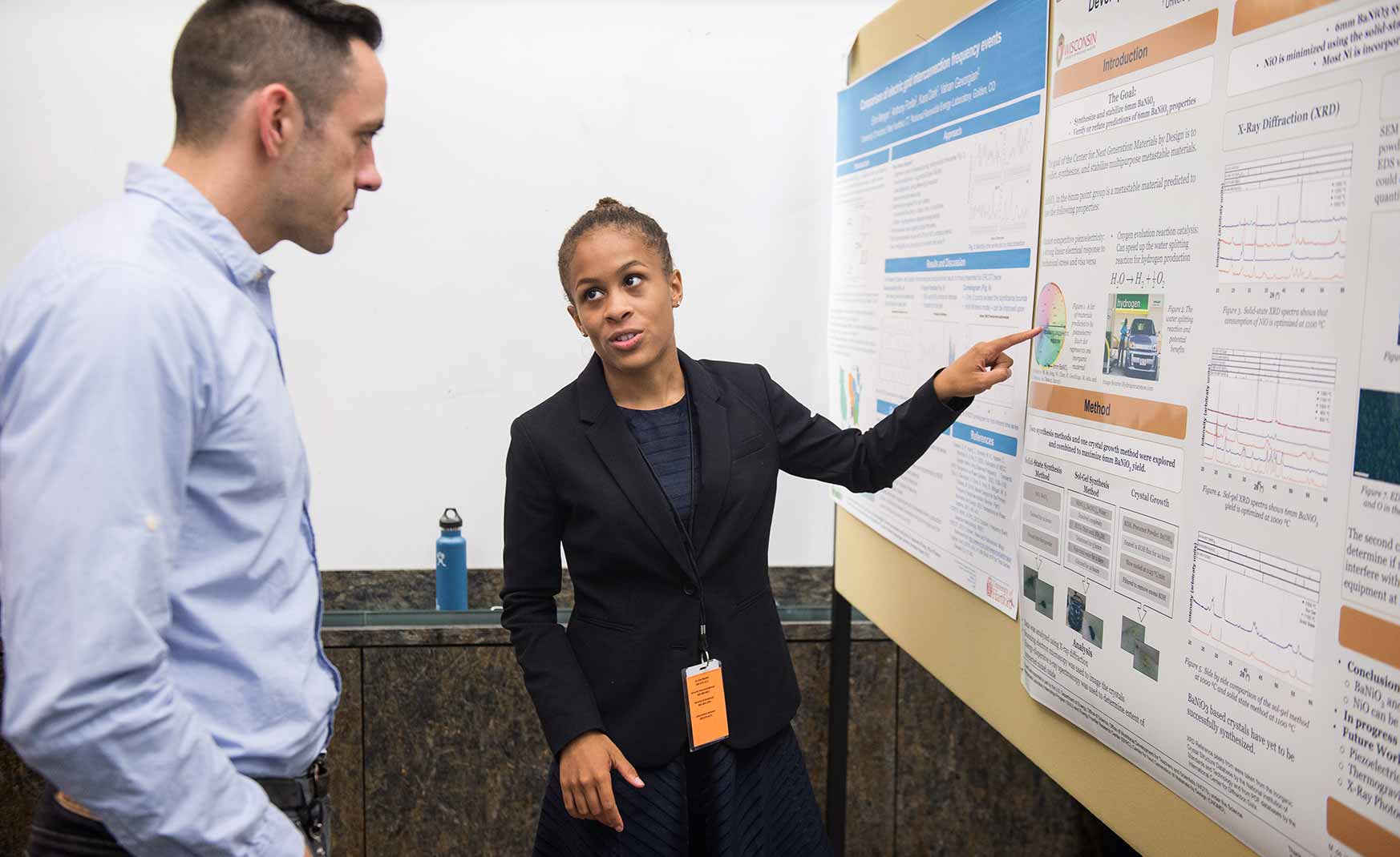 One person talks and points at a poster hanging on a board while another listens.
