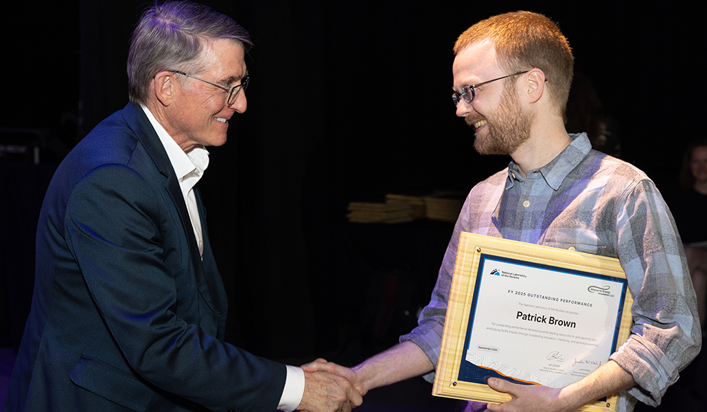 Two people shake hands and smile at one another as the one at right holds a plaque.