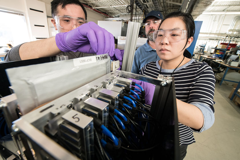 Three people in a laboratory handle objects while wearing gloves and protective eyewear.