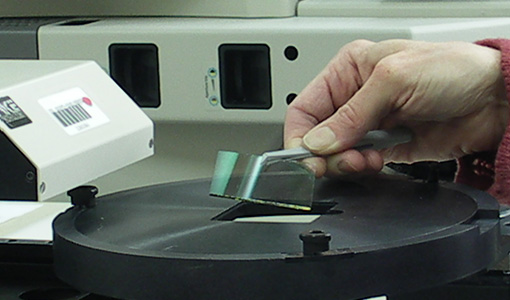 014485-flt Hand placing a small film or coated sample onto a circular testing stage of a laboratory instrument.