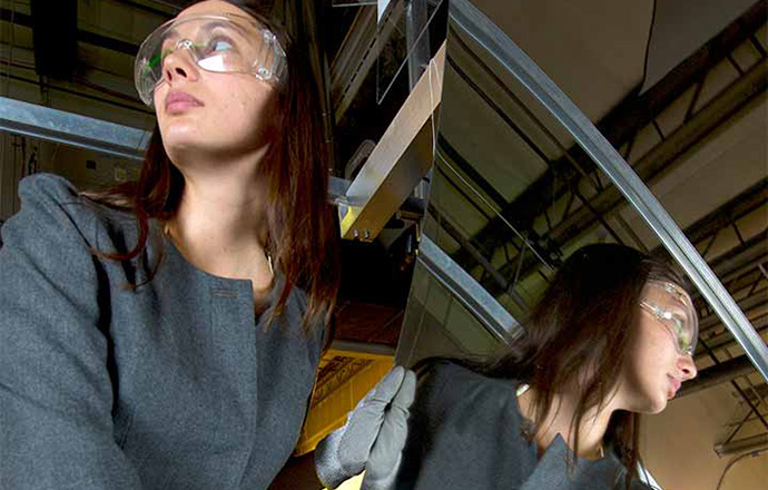 A woman in safety glasses is reflected in the large mirror she is moving with gloves.