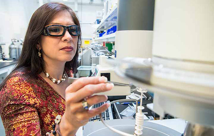 A woman in safety glasses reaches toward a piece of laboratoy equipment.