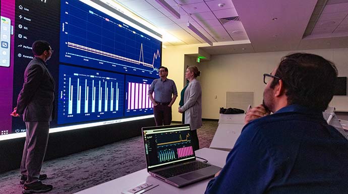 Researchers stand in front of large display screening discussing CySAT tool while another researcher sits in front of a laptop looking on.