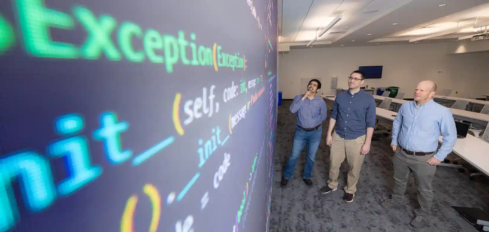 Three researchers stand and look up at cybersecurity data displayed on screen