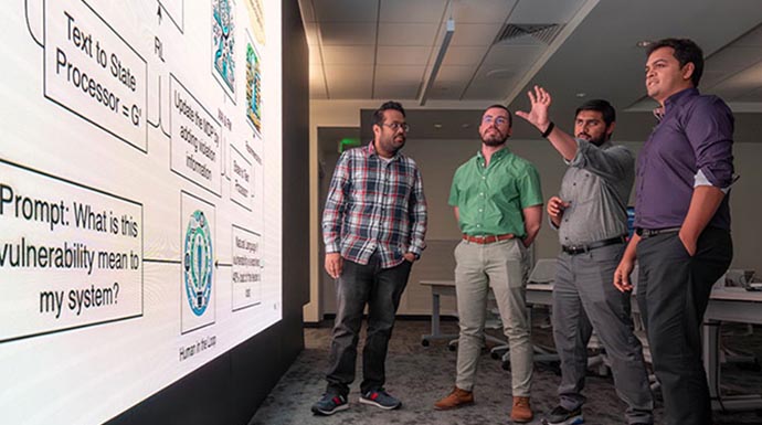 Four people stand and look at flow chart on large display screen.