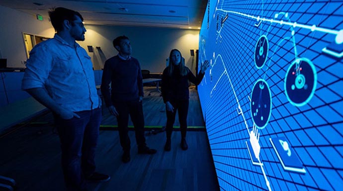 Three people stand in a dark room looking at a large wall display computer screen.