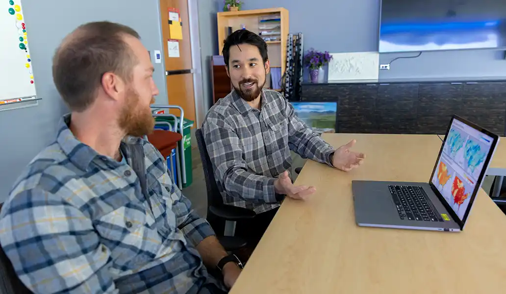 Two researchers work together on a laptop with maps on it