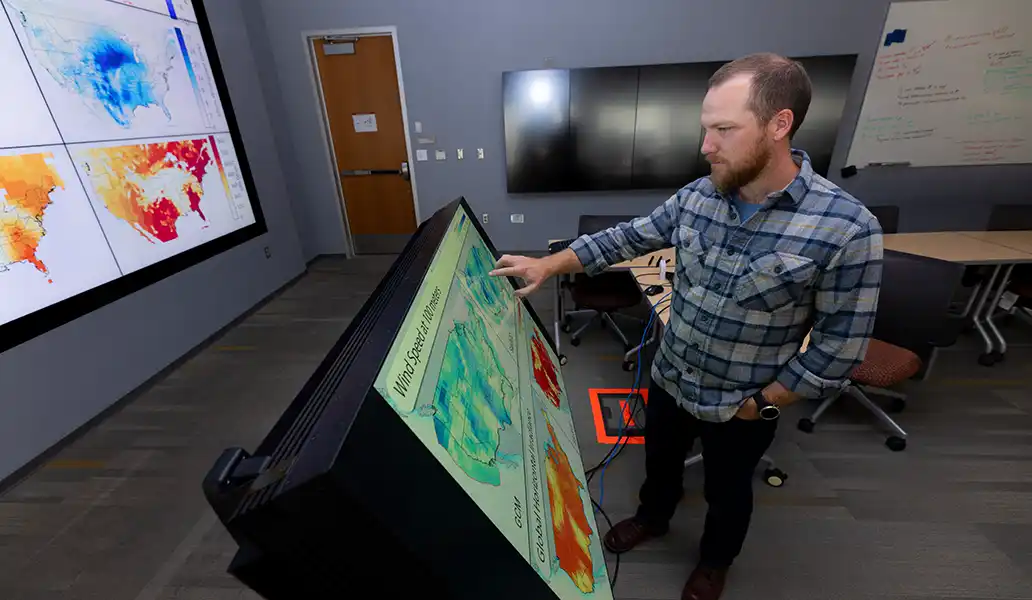 A researcher points at a wind speed map on a screen with additional maps displayed on a larger screen on the wall.