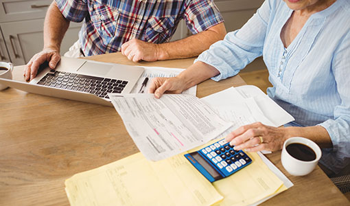 Two people look at a laptop and papers with a calculator and coffee mugs on the table.