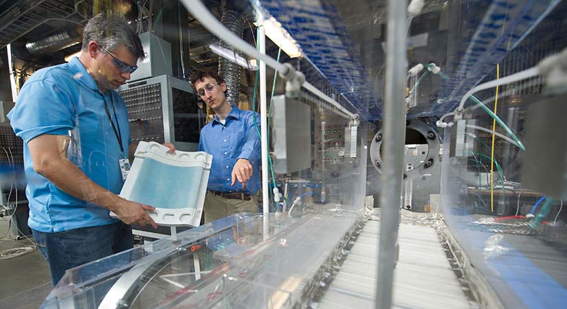 Two researchers communicate while standing on the side of a long piece of machinery.