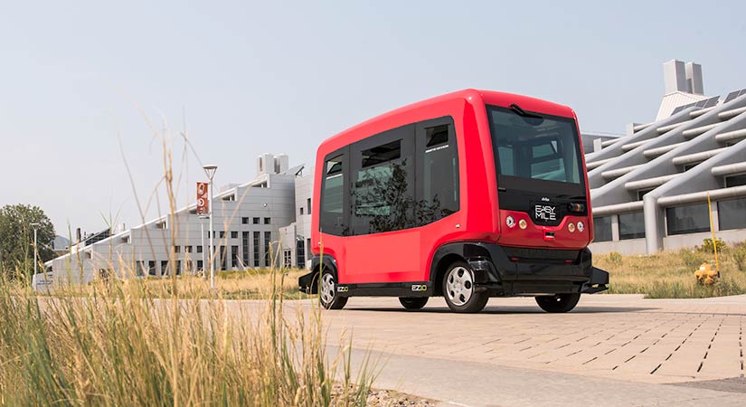 A photo of a red all-electric shuttle bus driving on a road by a large building.