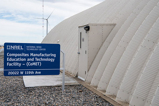 Photo of a white dome-shaped building with a sign that says, 'Composites Manufacturing Education and Technology Facility – (CoMET ),' and a wind turbine in the background.