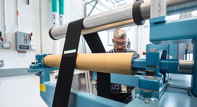 A photo of a man in a laboratory watching a black material run around various metal rolls.