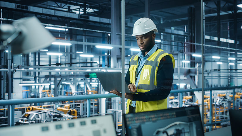 Worker stands inside a manufacturing plant overlooking laptop