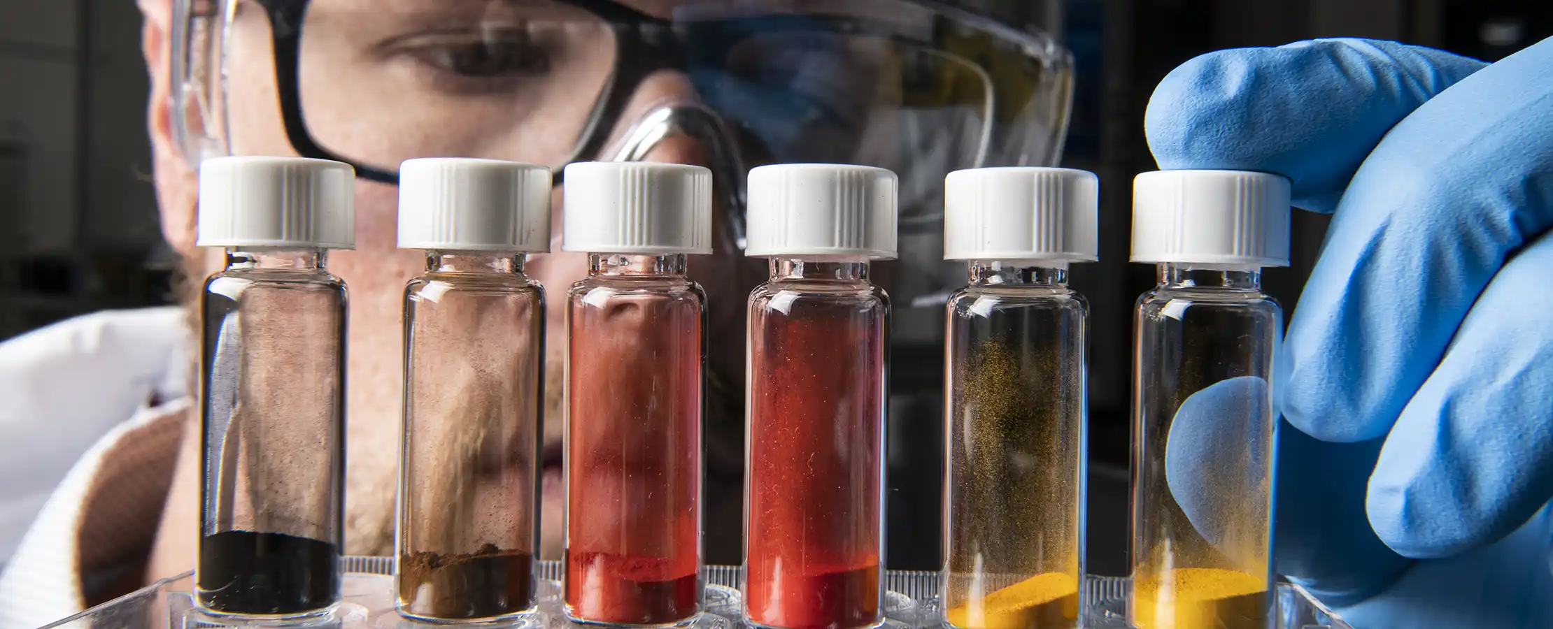 A researcher examines six vials containing different powdered materials.