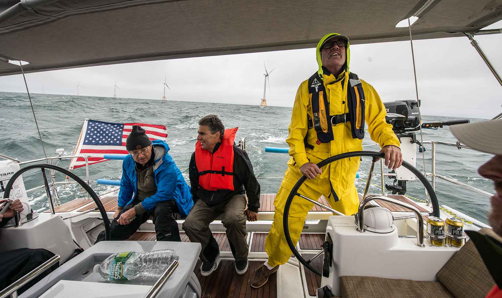 Three men on a boat in rough waters. Two in the background are looking at an offshore wind farm while one in the foreground is steering the boat.