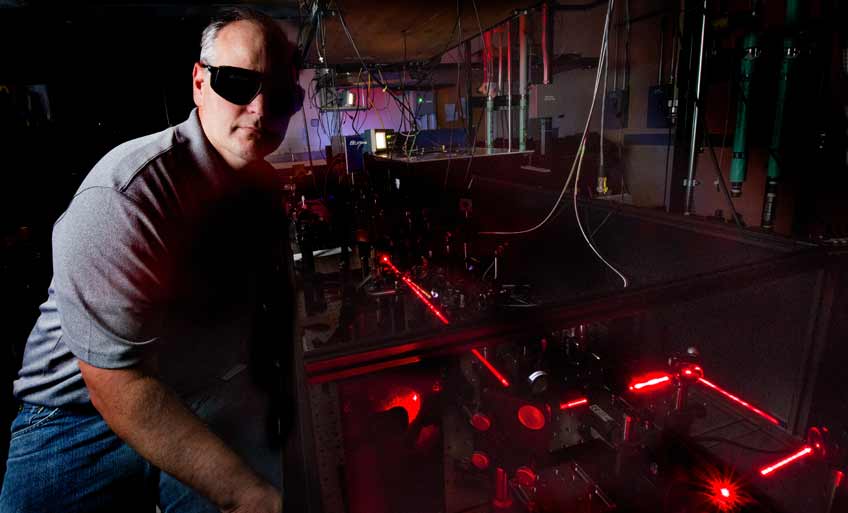 Photo of a man in a dark laboratory sitting next to a red laser beam.