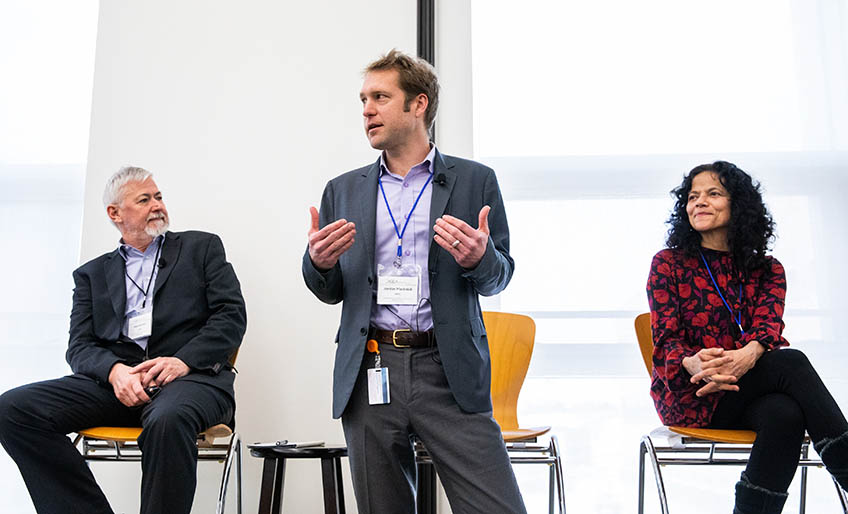A man stands to speak while two people listen on stage.