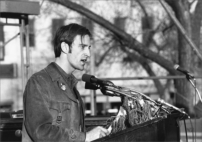 A man at an outdoor podium speaks into microphones