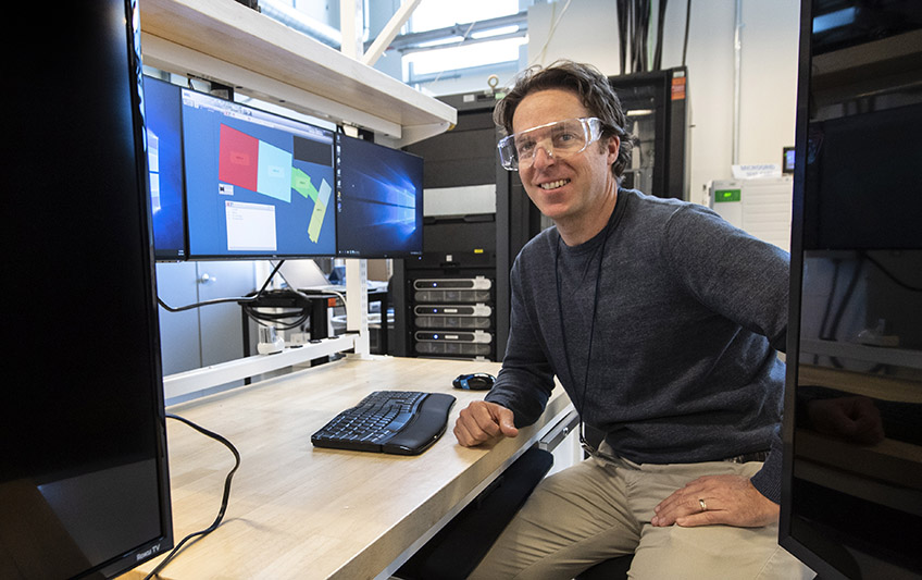 A man wearing safety glasses sits at a desk surrounded by computer monitors