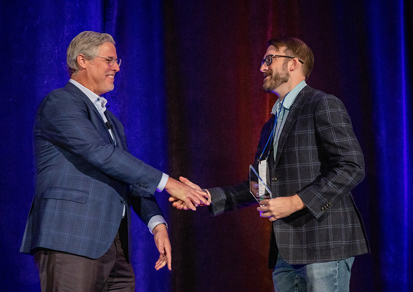Photo of two people shaking hands on an awards stage, with one person presenting an award to the other