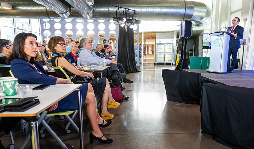 Photo of a person on stage speaking to a crowd of seated attendees in a large room
