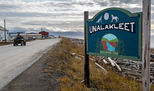 Continue reading about Sign says "Unalakleet Welcomes you" as a person rides an all-terrain vehicle down the road.