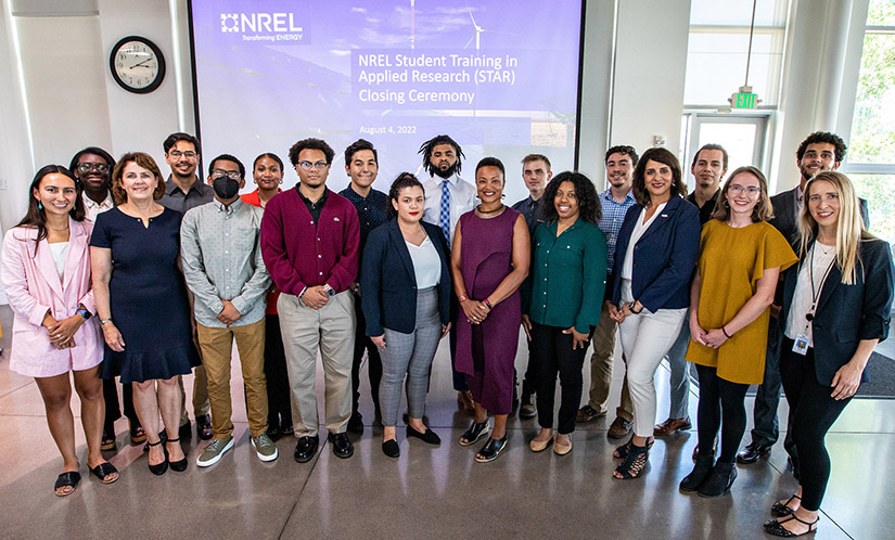 19 people standing and posing for group photo at NREL in front of a projected slide image that reads: