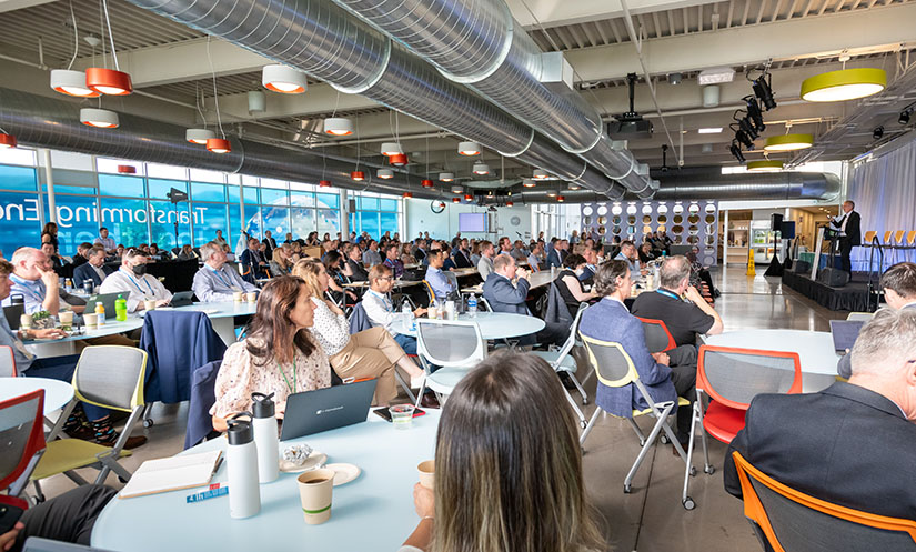 A large group of people sits around tables in a cafeteria while a person speaks at a podium.
