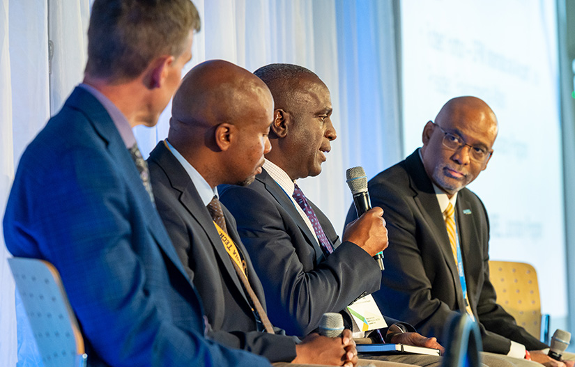 Three people on a panel watch while one person with a microphone speaks.