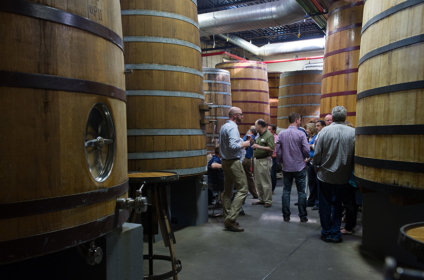People stand talking amidst huge beer brewing casks.