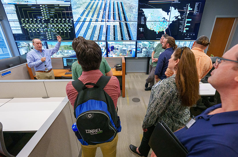 A group of people stand in front of a man talking with an image of solar panels on a screen next to him.
