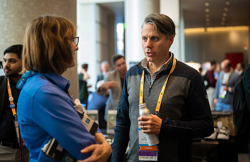 A man talks with a woman in a crowded exhibition hall with booths behind him.