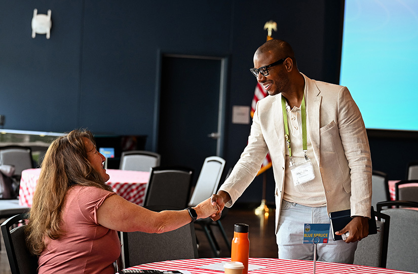 A man with glasses in a white blazer shakes the hand of a woman sitting at a table with a checkered tablecloth.