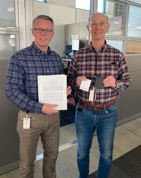 Two men stand in an office setting smiling at the camera. The man on the left holds a hard copy patent license agreement, while the other holds a prototype of his non-isocyanate polyurethane material.