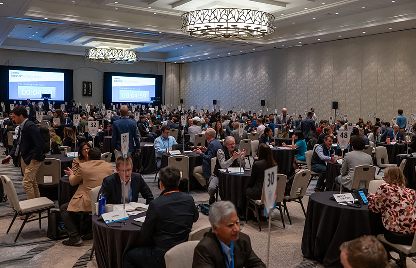 Hundreds of people sit at small tables in one-on-one meetings in a large ballroom.