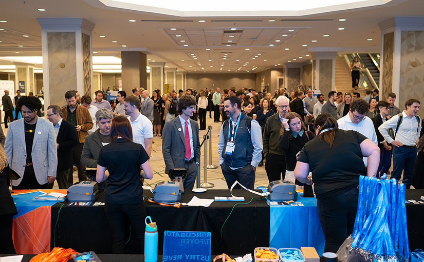 Two men talk with hundreds of people behind them in a large hotel area.