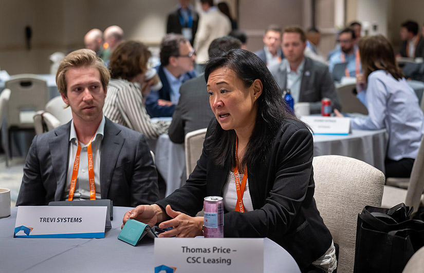 A woman sits at a round table next to a man speaking to someone off camera.