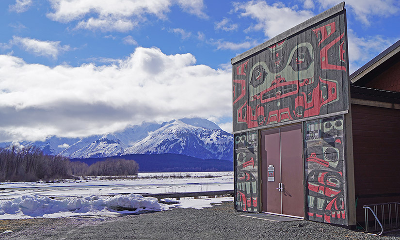 A building sits next to a snow-covered river with mountains in the background.
