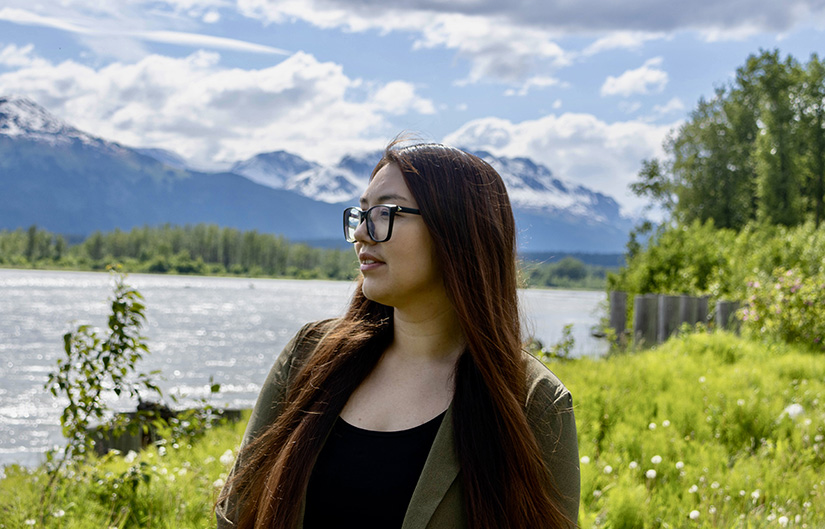 A person with long hair stands in front of a mountainous landscape.